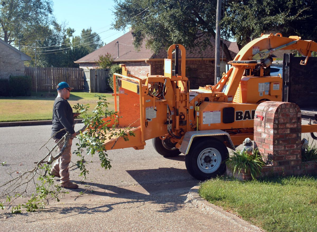 A worker feeding cut branches into a towable wood chipper parked on a suburban street in front of a brick house